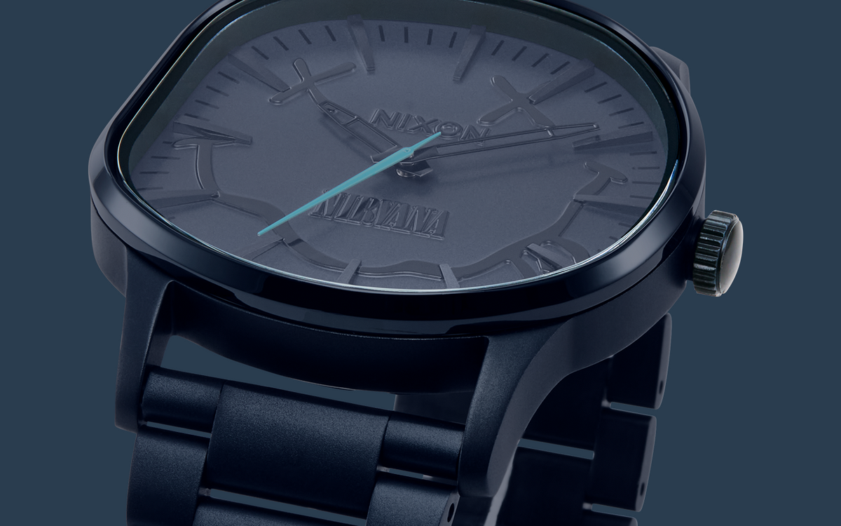 A close-up of a Nixon x Nirvana watch featuring a dark matte blue case, a matching dark dial with smiley face.