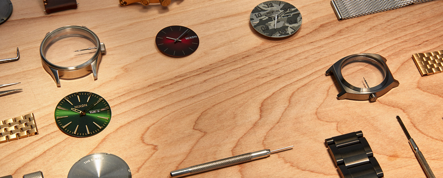 A selection of Nixon watch parts are spread out on a wooden table.