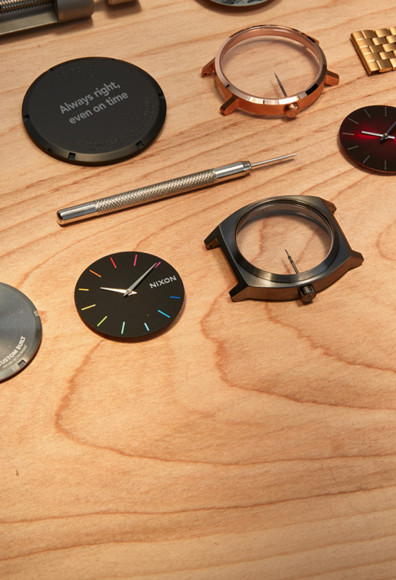 A selection of Nixon watch parts are spread out on a wooden table.