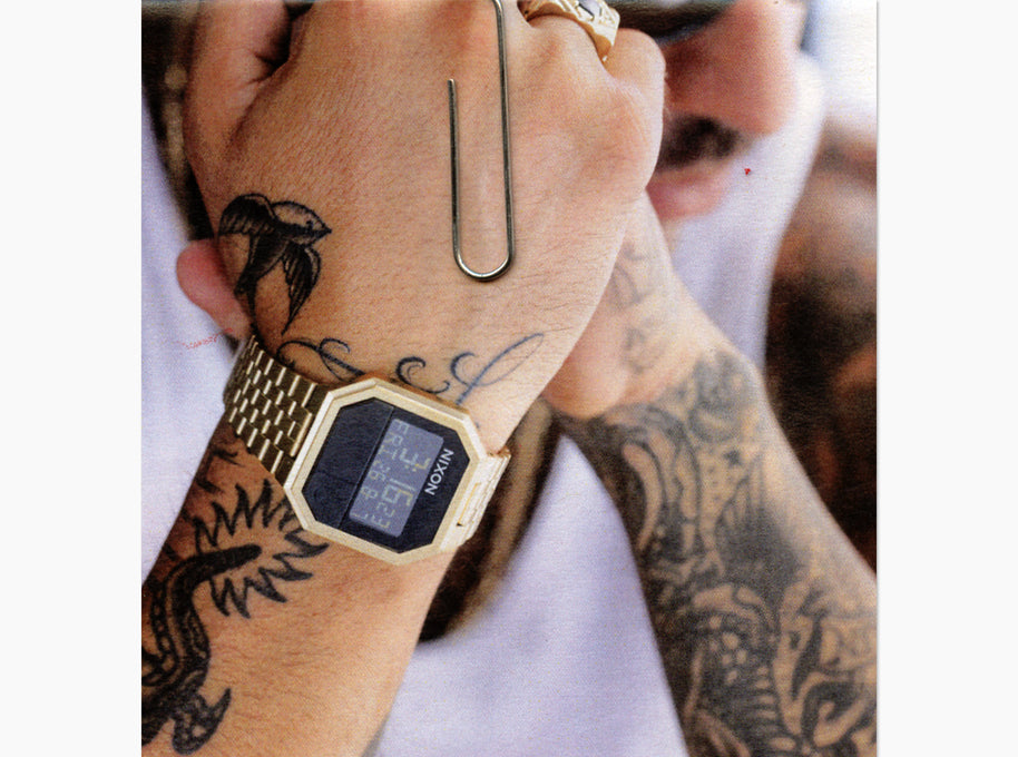 A close-up lifestyle shot of a person's tattooed wrist wearing a gold-tone digital watch with a gold link bracelet.