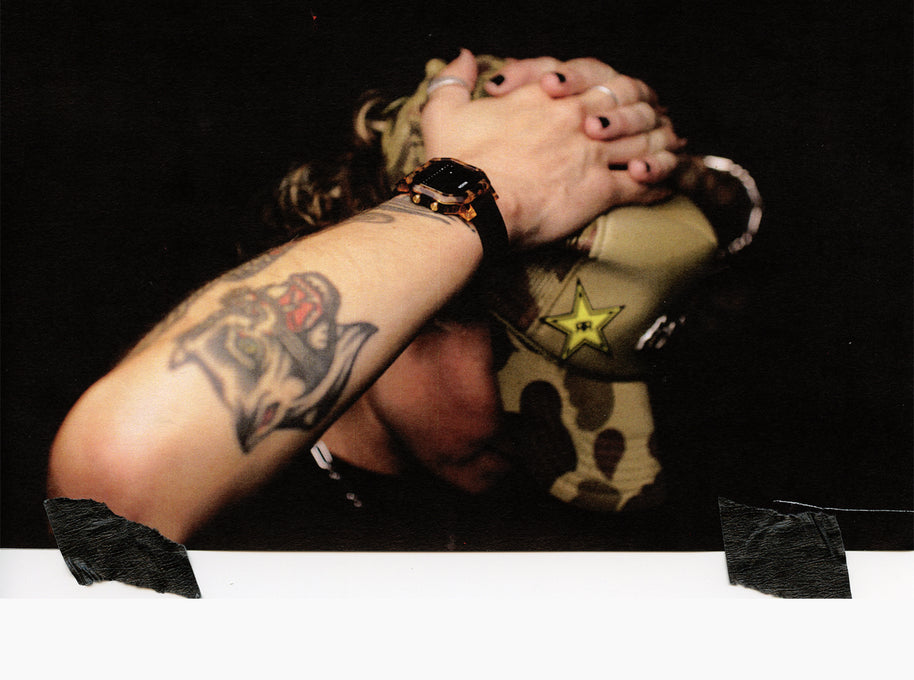 A close-up lifestyle shot of a person with tattoos covering their face, wearing a camouflage baseball cap with a yellow star logo and a black watch, set against a dark background.