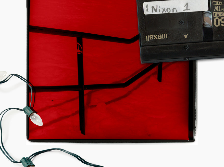 A square red photo, photographed on a white background. A black Maxell tape sits on the upper right corner of the artwork, and a string of Christmas lights is draped along the lower left edge.