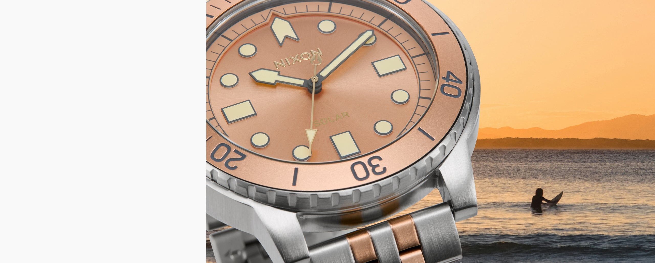 A close-up of a Nixon wristwatch with a rose gold dial and silver-toned metal case, set against a sunset beach scene with a surfer in the background.