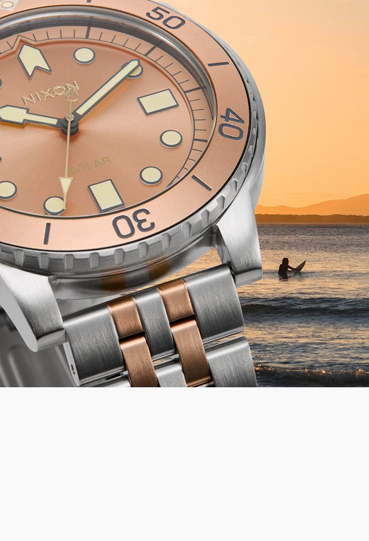 A close-up of a Nixon wristwatch with a rose gold dial and silver-toned metal case, set against a sunset beach scene with a surfer in the background.