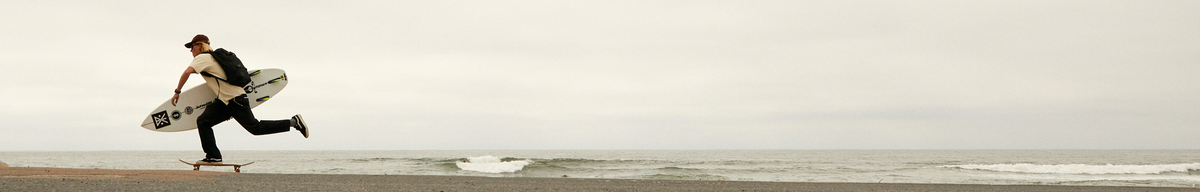 A person with a surfboard and backpack rides a skateboard along a beach with the ocean in the background.