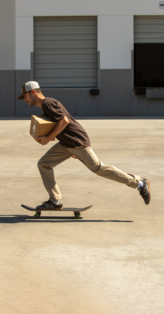 A man in a brown t-shirt and khaki pants rides a skateboard while carrying a cardboard box in an outdoor industrial lot.