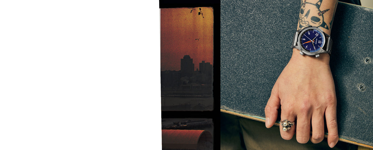 A close-up lifestyle shot of a man's hand and wrist wearing a silver-toned wristwatch with a blue dial, resting on a dark gray textured surface next to a skateboard. The background is split between a solid white area on the left and a warm-toned cityscape silhouette on the right.