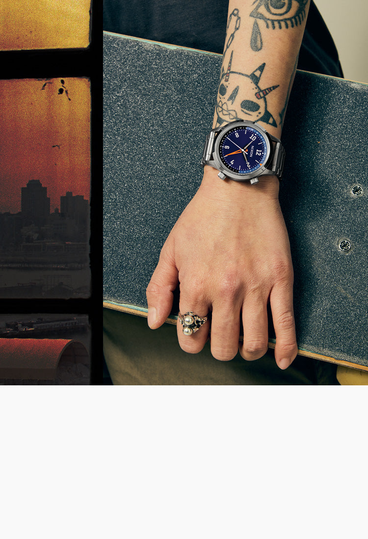 A close-up lifestyle shot of a person's tattooed arm and hand resting on a skateboard, wearing a silver-toned wristwatch with a deep blue dial and a silver ring on the finger.