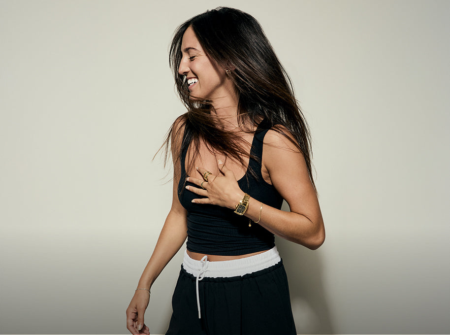 Woman wearing a black tank top and black pants against a plain background wearing a Nixon watch